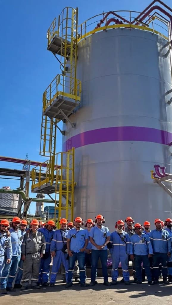 Equipe com EPI em frente a tanque industrial no Projeto Sucuriú, sob céu azul